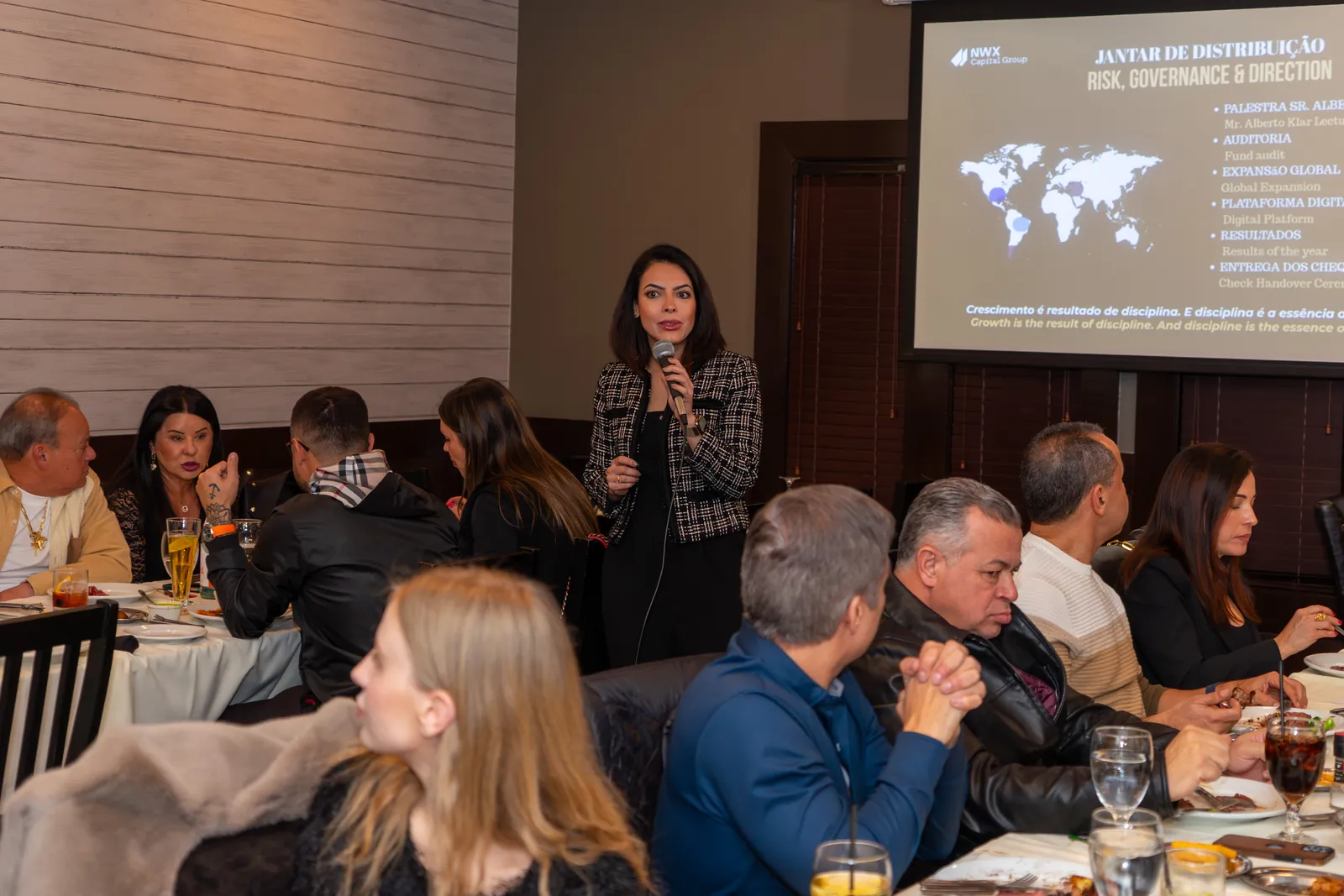A speaker with a microphone addressing a seated dinner, a presentation slide behind her