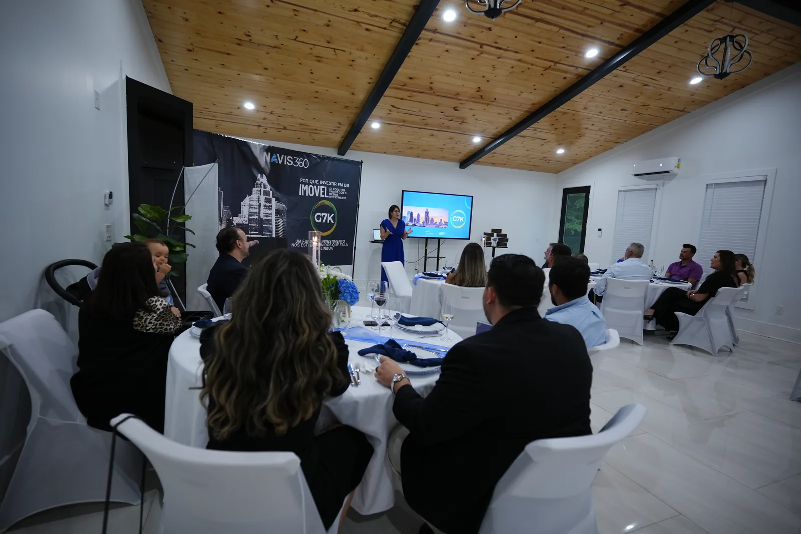 A presentation in a meeting room, people seated at round tables facing a screen
